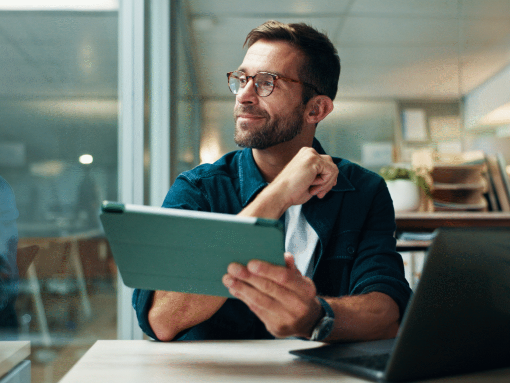 man at a desk with a laptop in a office holding a tablet and smiling while thinking about adaptive learning systems